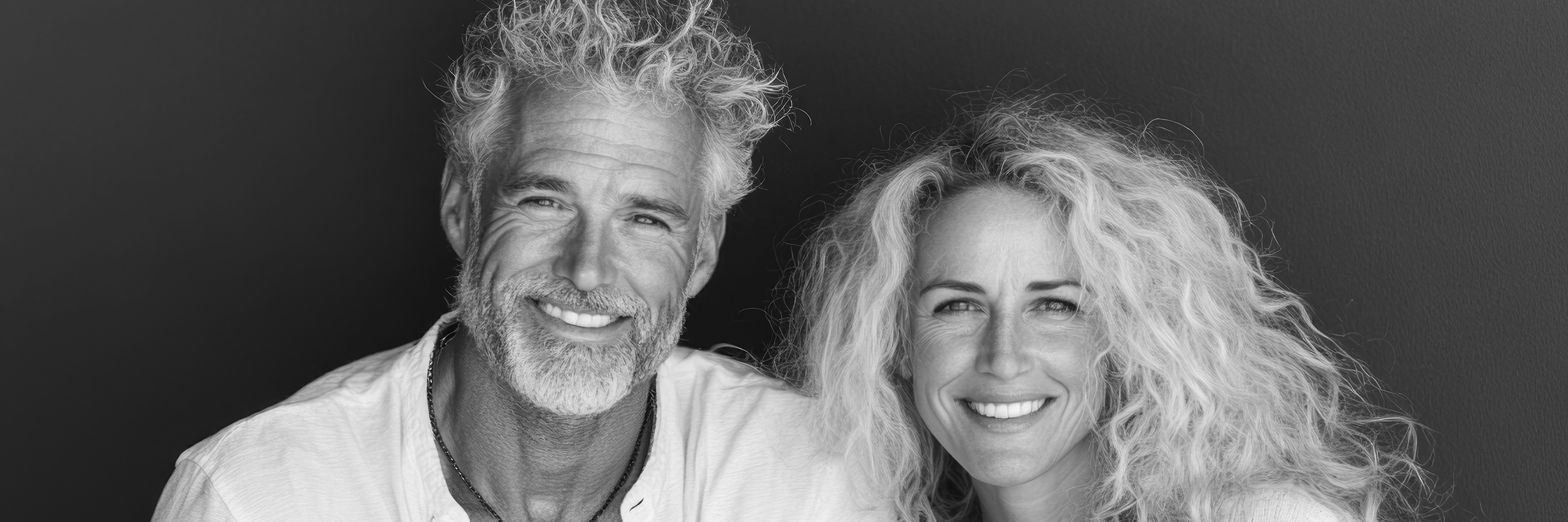 Black and white photo of an older man and woman smiling, with curly hair, against a dark background.