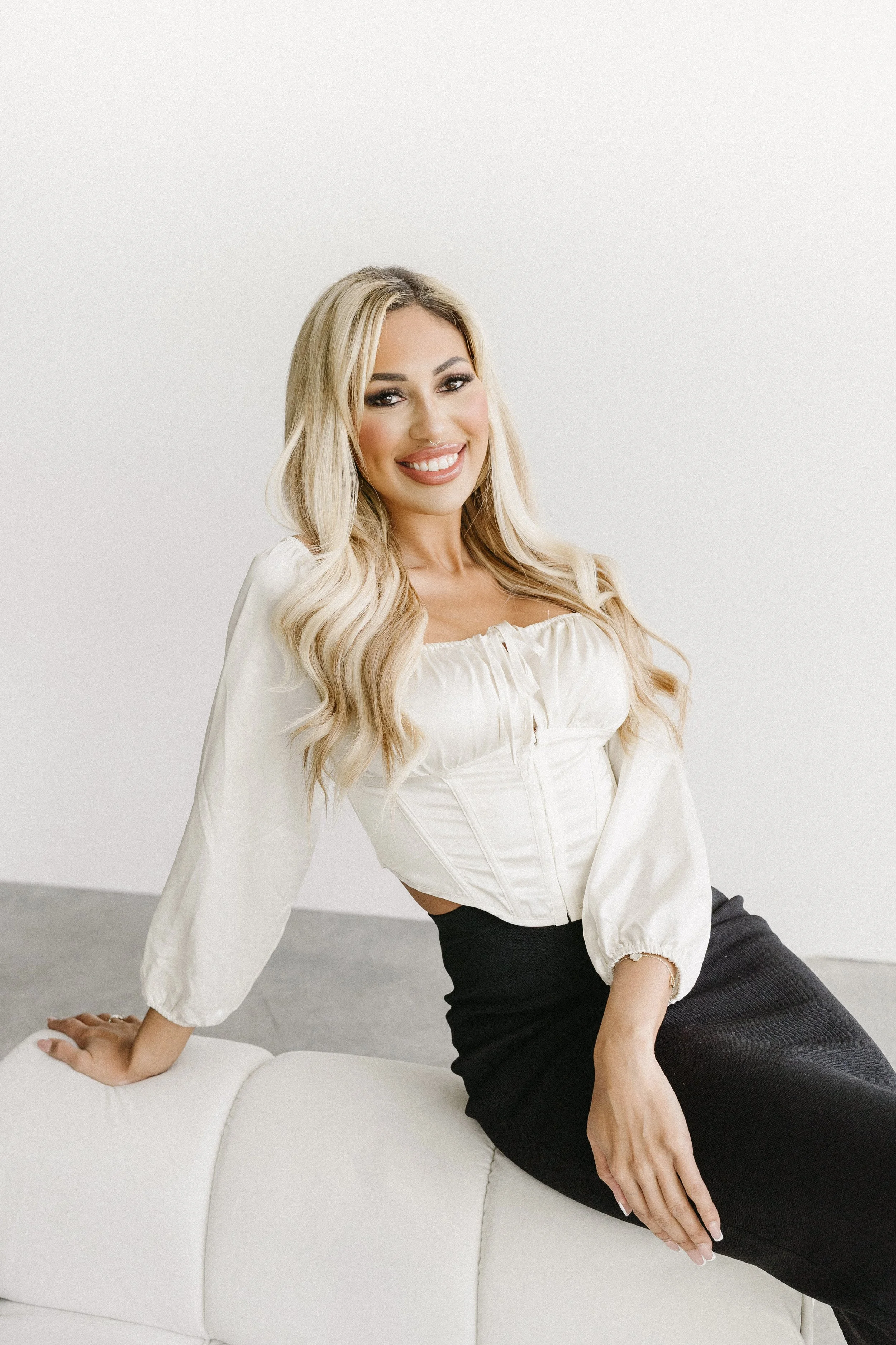 A woman with long blonde hair, wearing a white blouse and black skirt, sitting on a white couch, smiling.