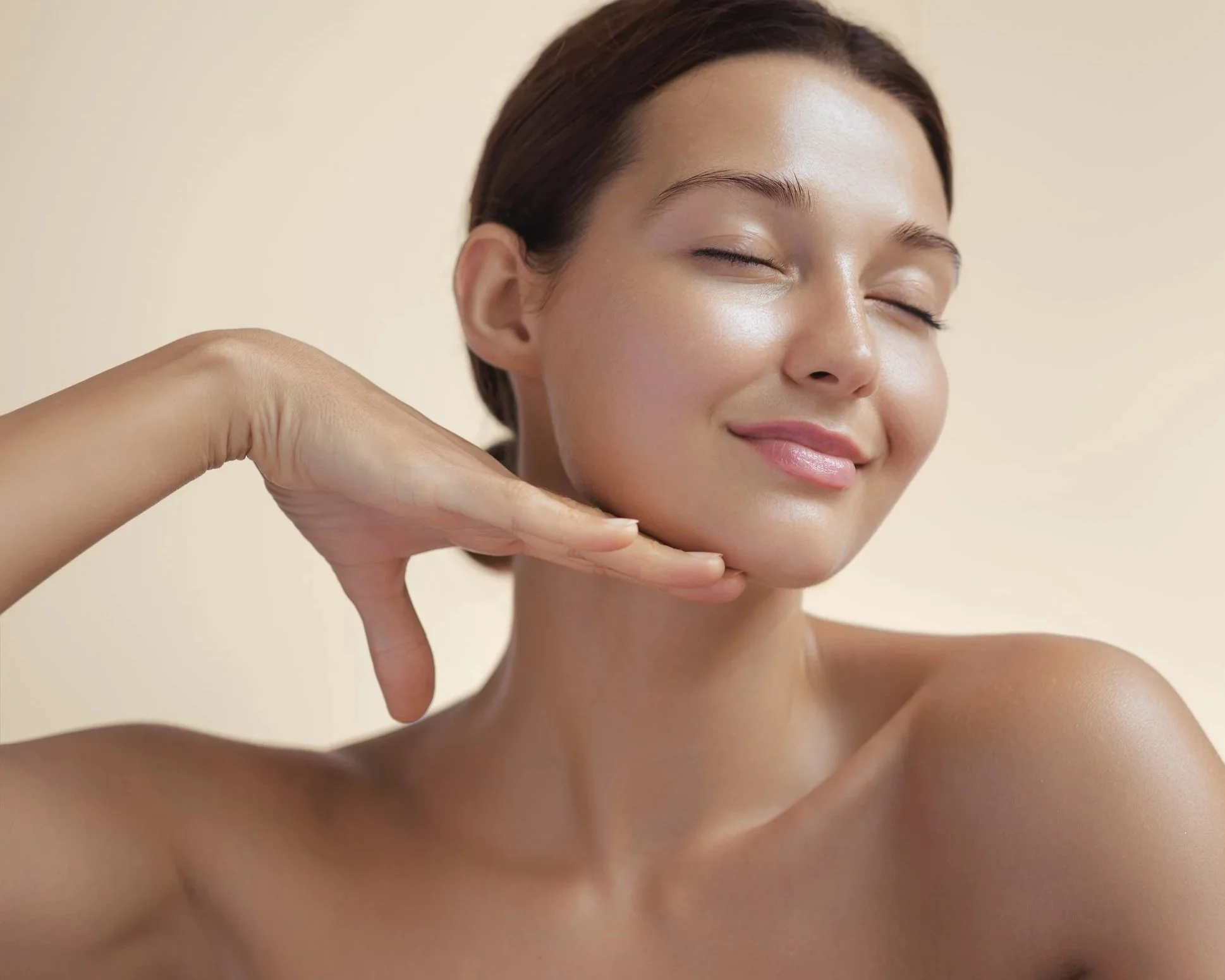 Close-up of a smiling woman with clear, glowing skin, resting her chin on her hand, with bare shoulders against a neutral background.