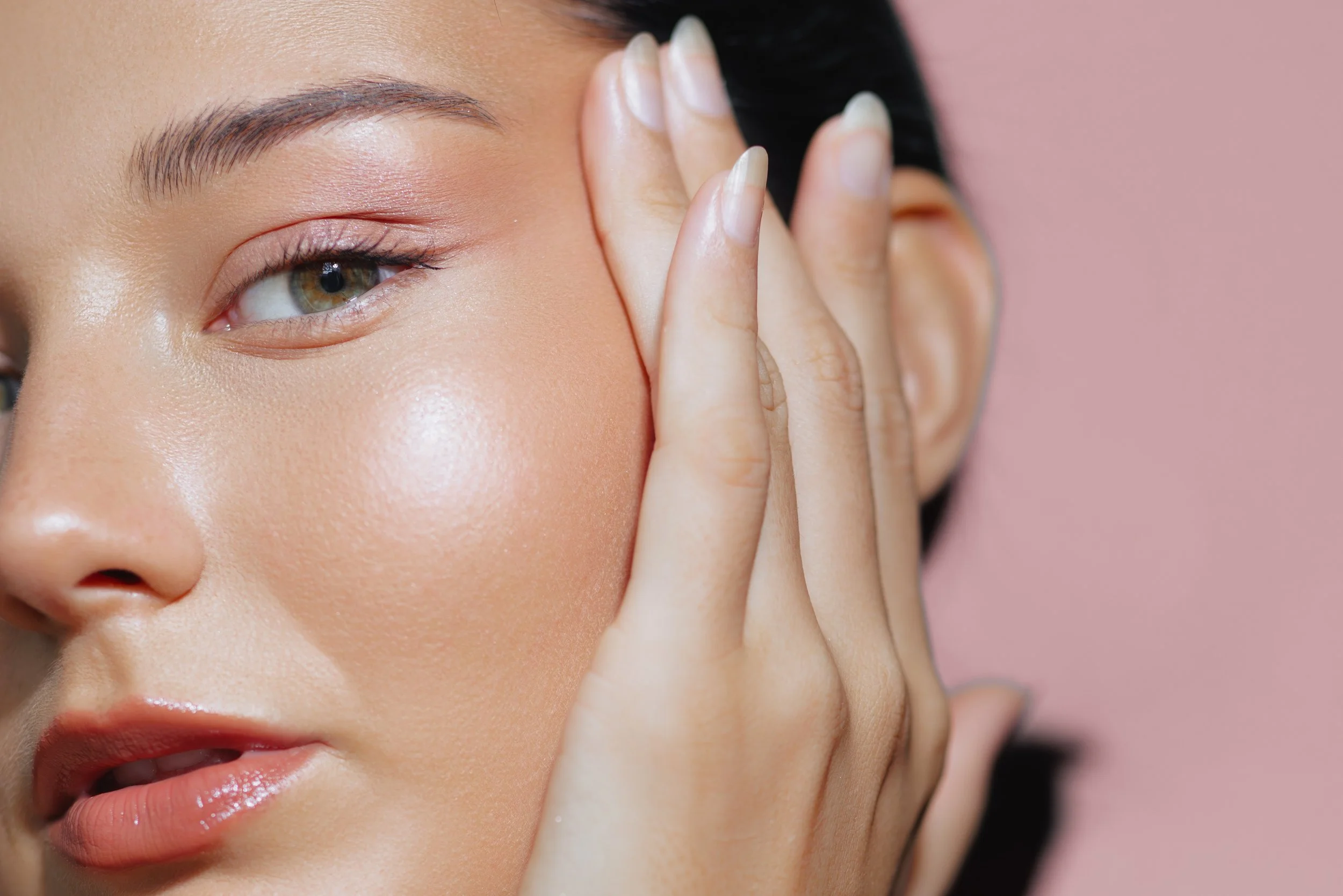 Close-up of a woman with flawless skin, wearing natural makeup, gently touching her face with her hand, against a soft pink background.
