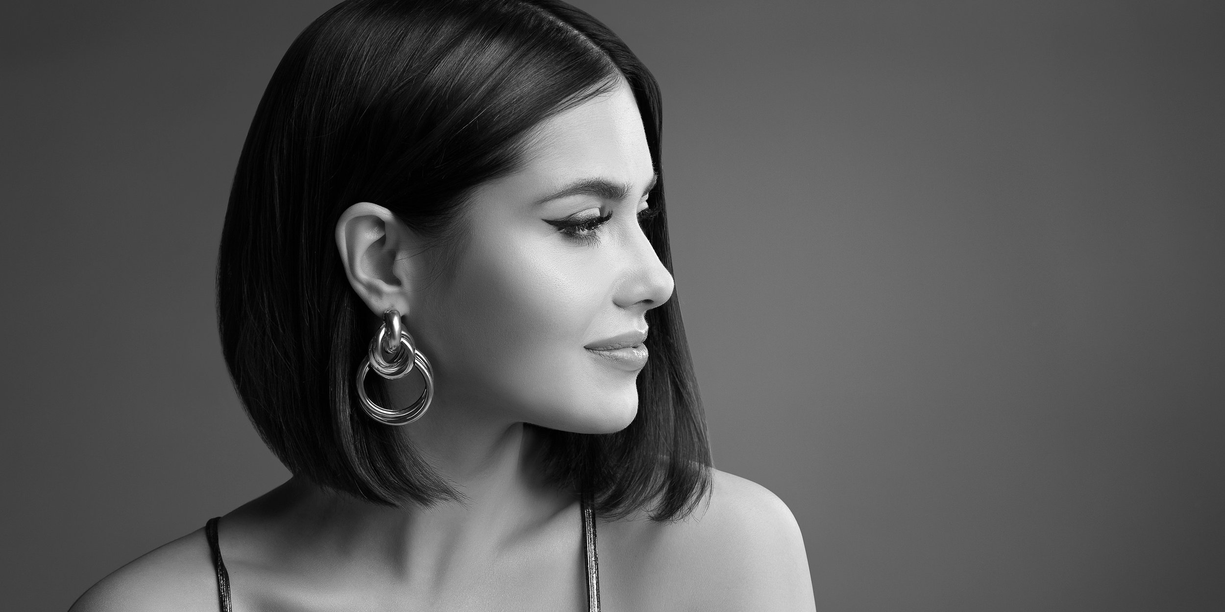 Black and white portrait of a woman with straight, shoulder-length hair, wearing large hoop earrings and a thin strap top, looking to the right with a subtle smile.