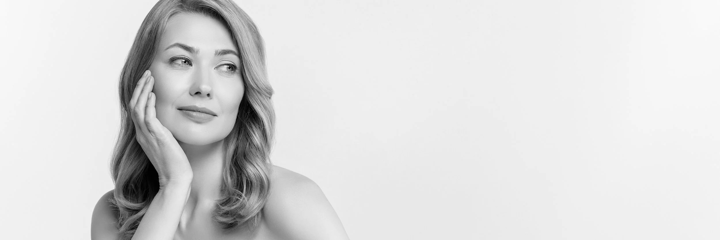 A black-and-white photo of a woman with wavy hair touching her face gently, looking slightly to the side with a calm expression, against a plain light background.