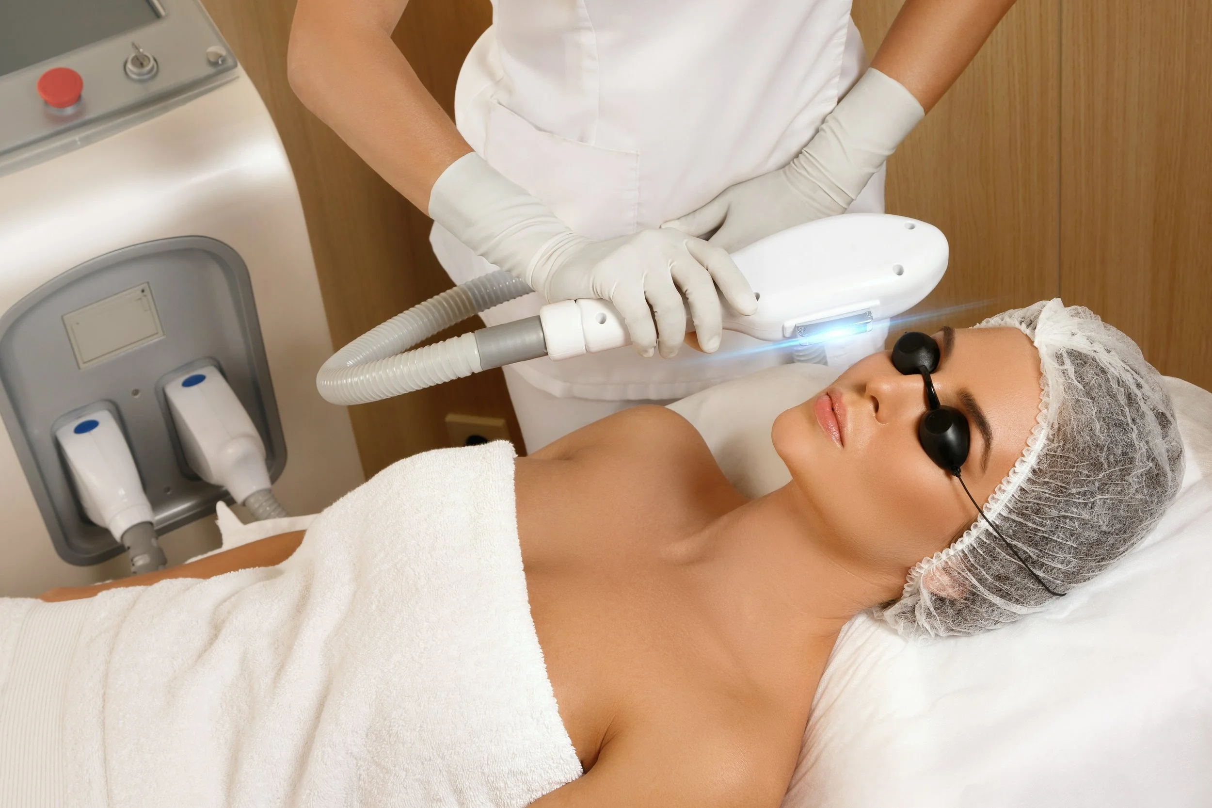 A woman lying on a medical bed, wearing protective goggles and a hairnet, receives laser treatment from a healthcare professional wearing gloves, white uniform, and holding a laser device.