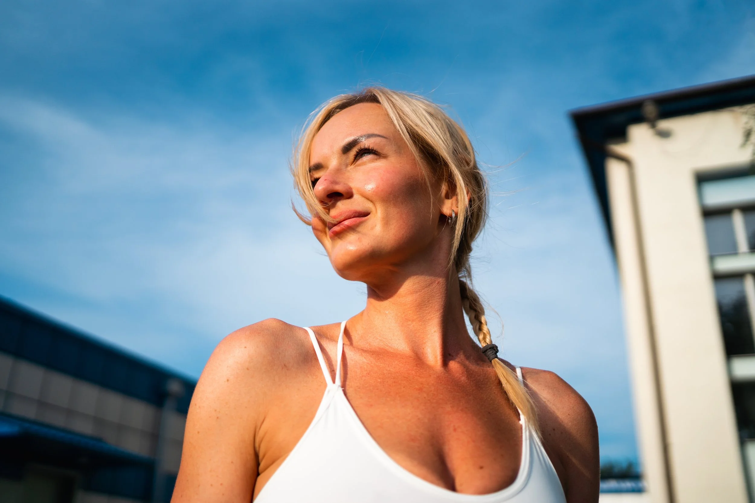A woman with blonde hair in a braid wearing a white tank top smiling outdoors during daytime, with modern buildings and a blue sky in the background.