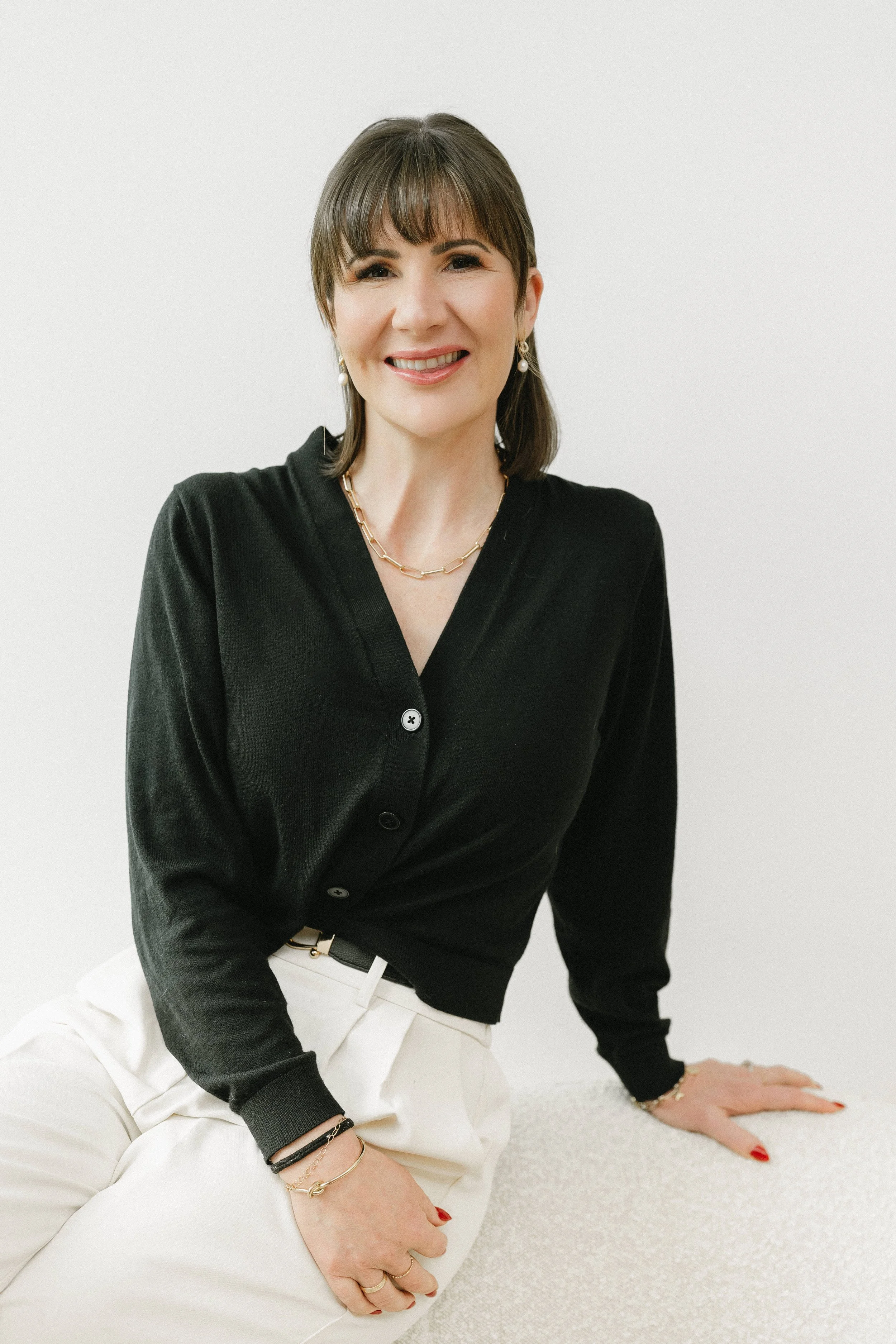 A woman smiling, wearing a black cardigan, white pants, and jewelry, sitting on a light-colored surface against a white background.
