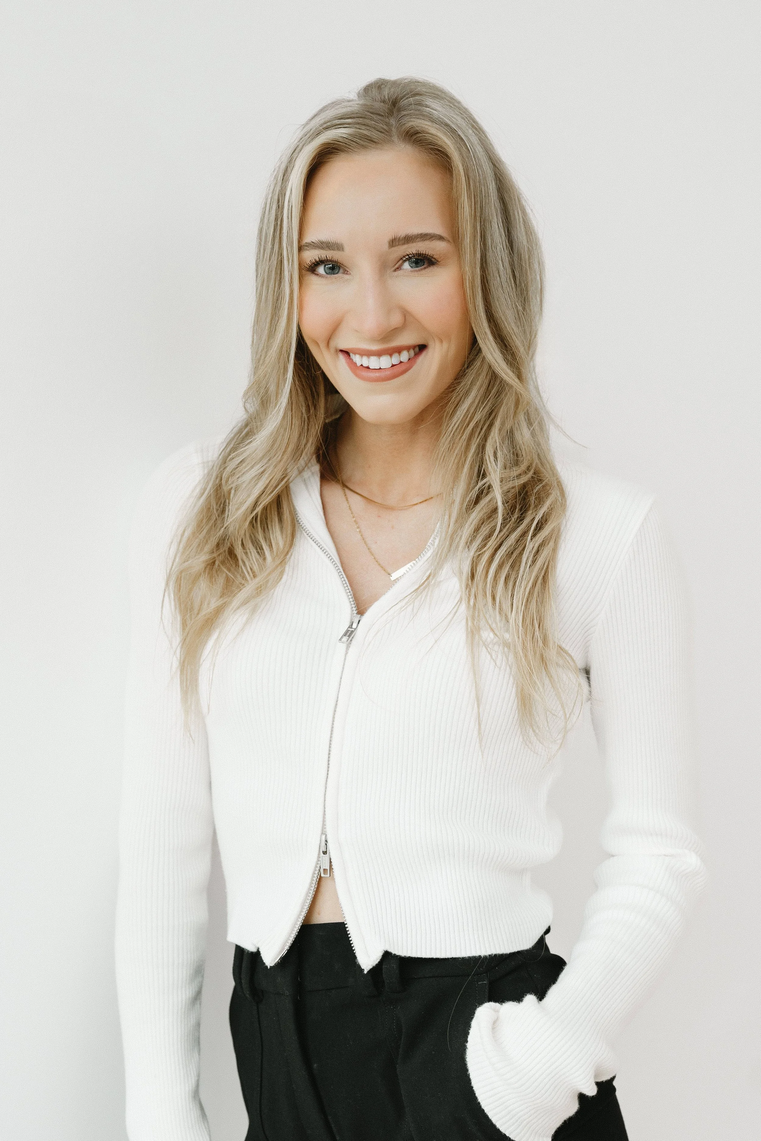 A young woman with long blonde hair, smiling, wearing a white zip-up sweater and black pants, against a plain white background.