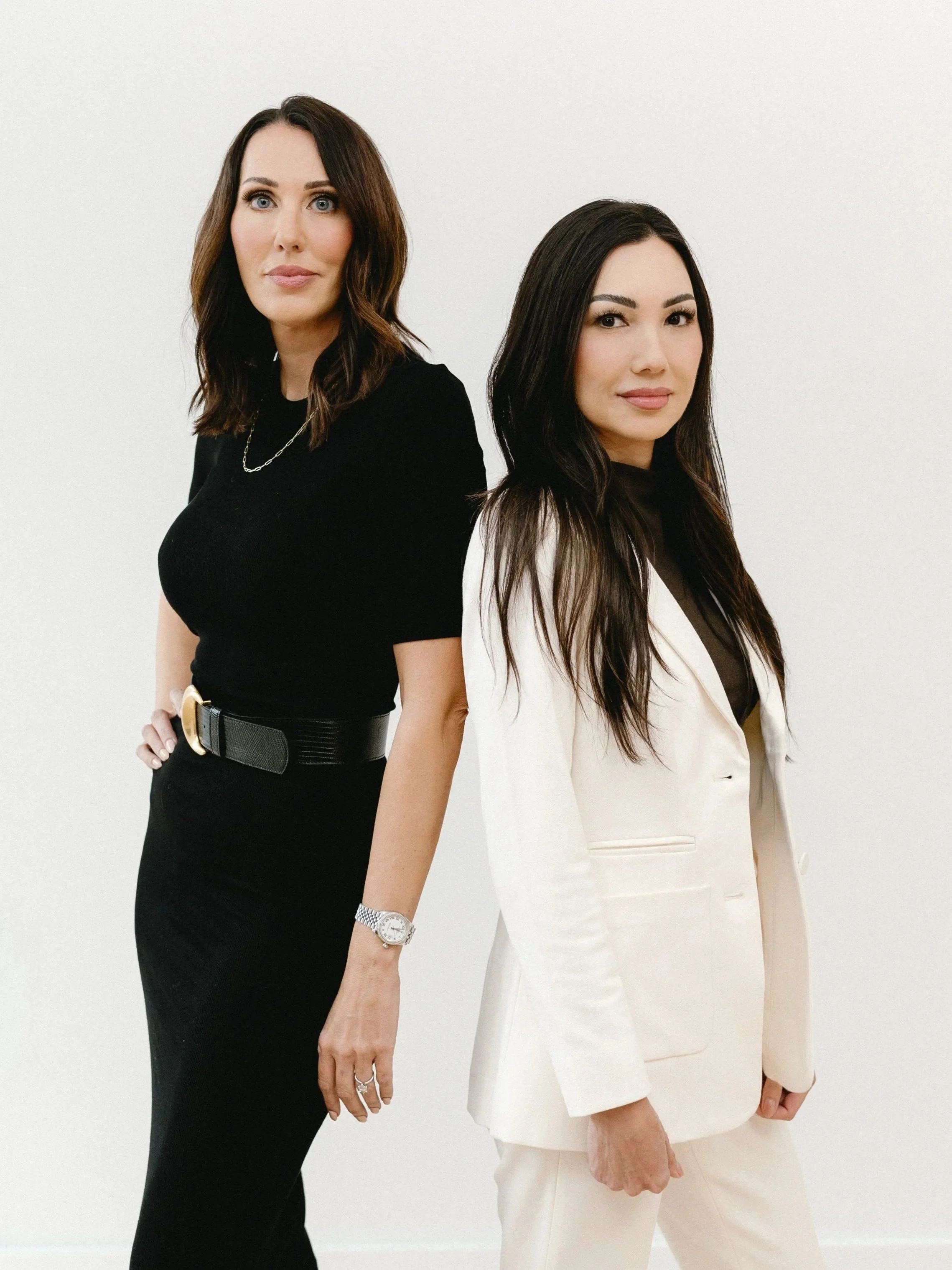 Two women stand side by side against a white background, looking at the camera. The woman on the left wears a black dress with a wide belt, has shoulder-length brown hair, and wears jewelry. The woman on the right wears a white suit with a black top, has long black hair, and a subtle smile.